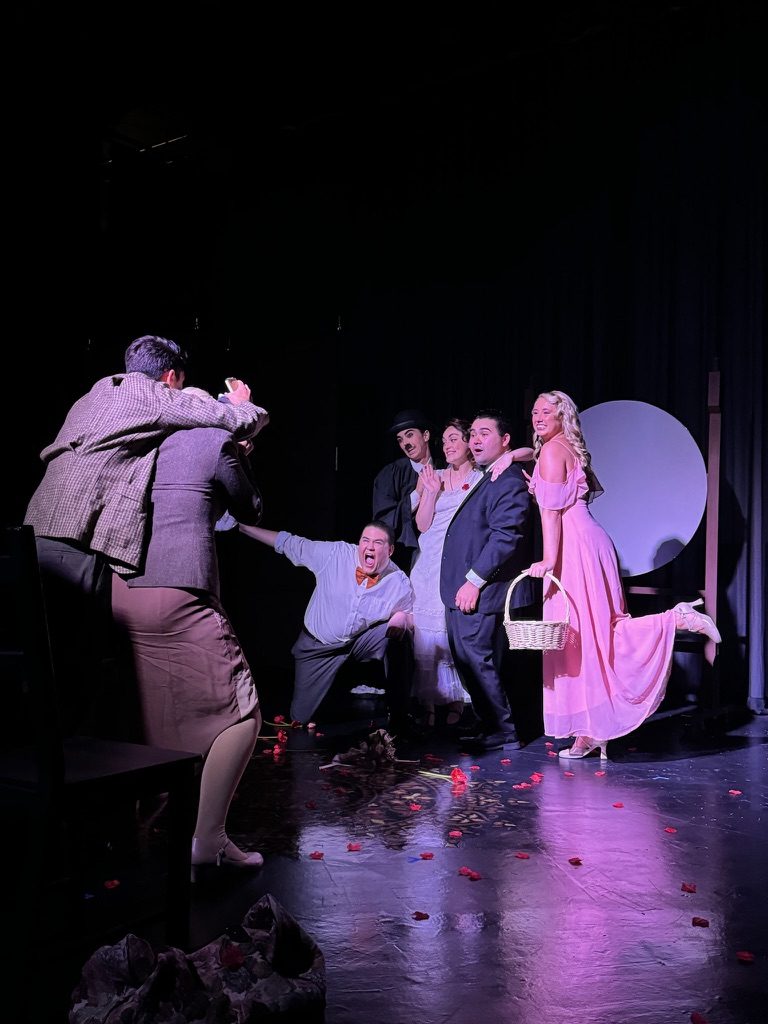 On stage: a photographer crouches before a posed group in 1915 dress