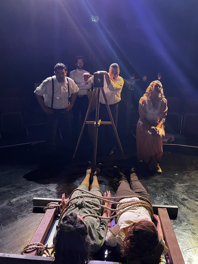 On stage: two figures tied to railroad tracks while a crew films